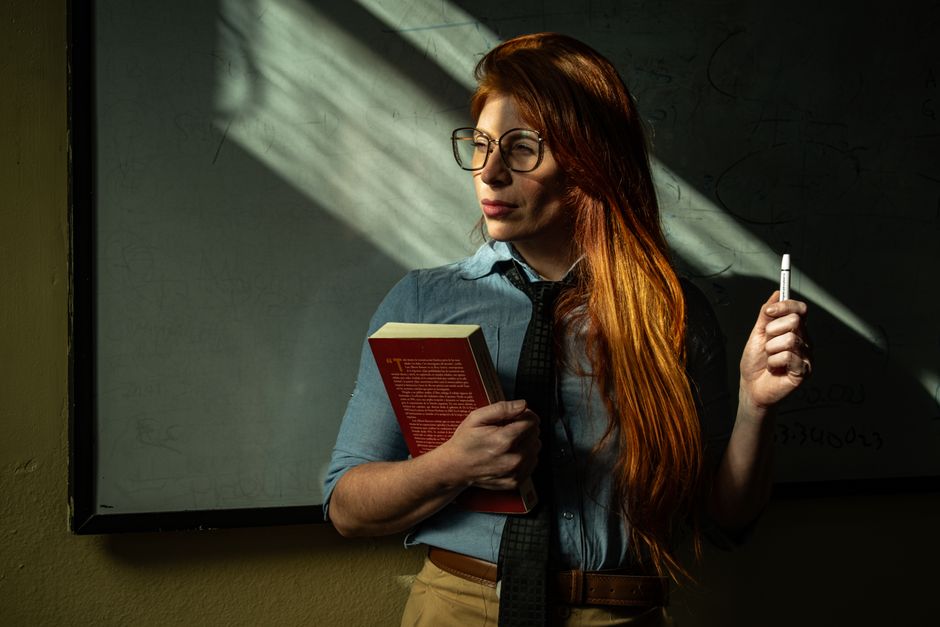 Thoughtful teacher standing by chalkboard holding book and marker, immersed in sunlight.